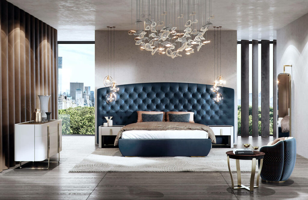 A luxurious bedroom featuring a stunning luxury chandelier, a tufted navy blue headboard, modern furniture, and large windows with city views.