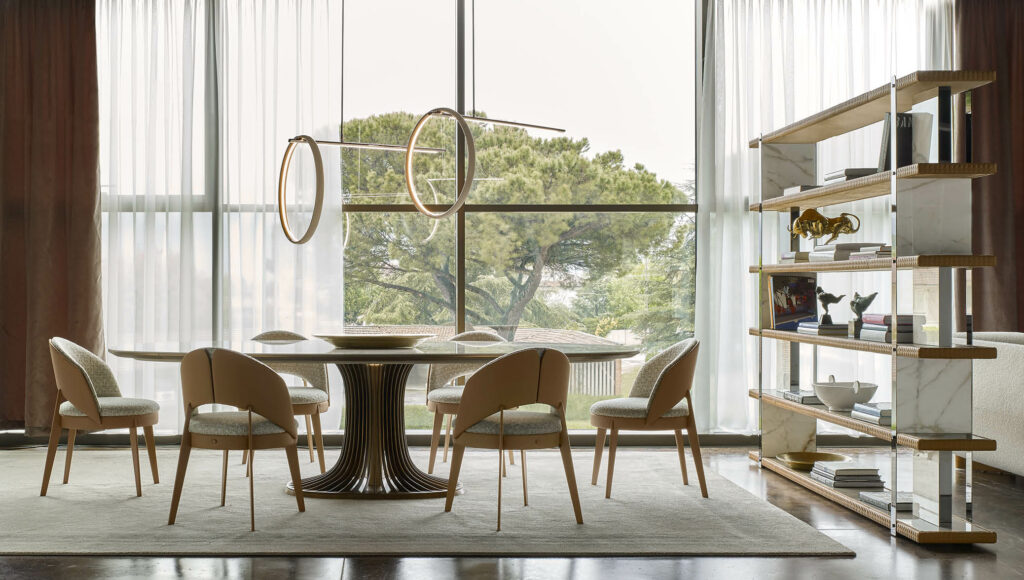 A modern dining room with a large window, a round table with beige chairs, and a luxury chandelier. A marble and wood bookshelf is on the right.
